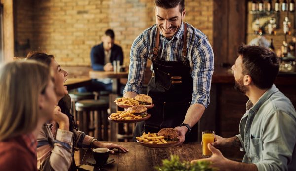 Group of happy friends having fun while waiter is serving them food in a pub. Focus is on waiter.